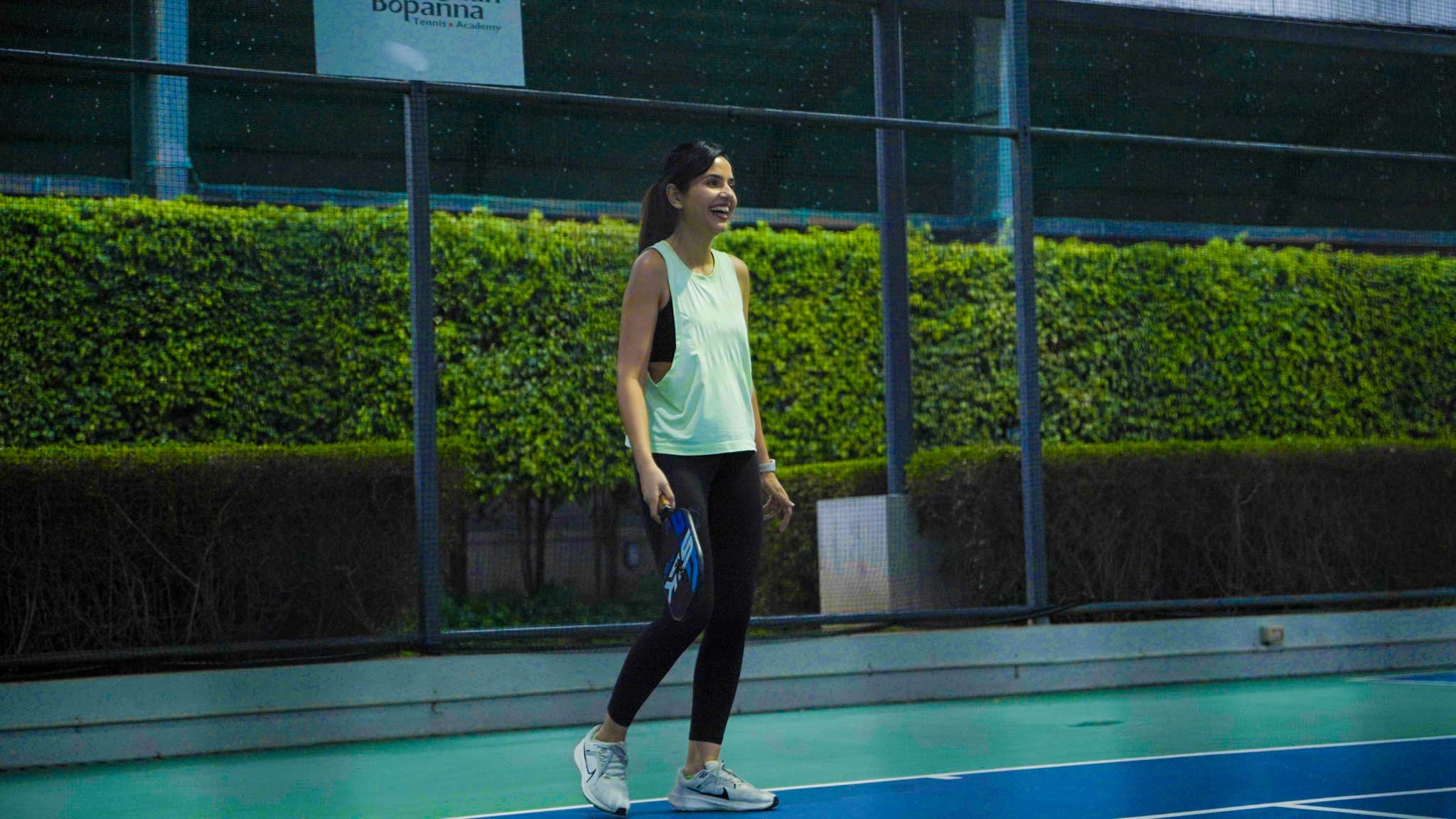Young woman smiling and holding a pickleball paddle on a court with a security guard in the background.